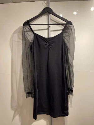 Svart klänning med meshärmar - Svart kort klänning med transparenta meshärmar och lätt rynk vid bysten. Klänningen har en rak passform och långa ärmar i tunn mesh som ger en cool och trendig look. Perfekt för dig som gillar stilrena plagg med en twist.