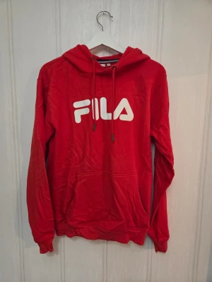 Röd hoodie från FILA med logga - Säljer en röd hoodie från FILA med stor vit logga på bröstet. Tröjan har huva med snörning, lång ärm och en klassisk magficka framtill.