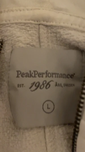 Vit teddyjacka Peak Performance L - Mysig vit teddyjacka från Peak Performance med broderad logga och fjäderdetalj på bröstet. Jackan har dragkedja och är tillverkad i ett fluffigt teddy-material som håller dig varm. Perfekt för dig som gillar en clean och stilren look.Villig att sänka pris!