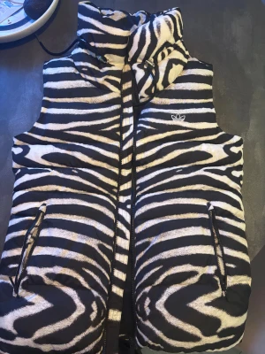 Adidas dunväst med zebramönster - Snygg dunväst från Adidas med svartvitt zebramönster och hög krage. Västen har dragkedja framtill, två fickor med dragkedja och Adidas logga på bröstet. Perfekt statement-plagg för dig som vill sticka ut.