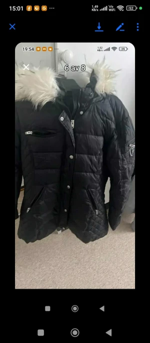 Svart lång pufferjacka med päls - Svart pufferjacka med lång passform och snyggt quiltad design. Jackan har en huva med fluffig päls, flera dragkedjeförsedda fickor och en cool patch på ärmen. Perfekt för kalla dagar och har en modern look som funkar till streetstyle.