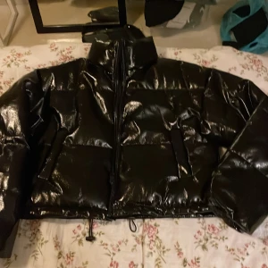 Svart glansig pufferjacka H&M Divided XS - Svart pufferjacka från H&M Divided i storlek XS. Jackan har en glansig finish, hög krage och dragkedja framtill. Tillverkad i polyester med fyllning och coating som ger en cool, modern look. Perfekt för dig som vill sticka ut med en edgy stil.Kort Midje modell.
