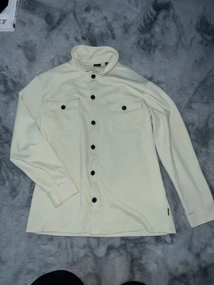Beige overshirt med svarta knappar - Storlek M funkar även S Snygg beige overshirt med svarta knappar och två bröstfickor med lock. Skjortan har lång ärm och en mjuk, lite tjockare bomullskänsla. Perfekt att slänga över en t-shirt för en chill look. Klassisk krage och stilren design.