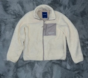 Beige teddyjacka från Jack & Jones M - Mysig beige teddyjacka från Jack & Jones Originals Studio med hög krage, hel dragkedja och en grå bröstficka med dragkedja. Jackan har två sidofickor och är tillverkad i mjuk fleece som håller dig varm under kyliga dagar.