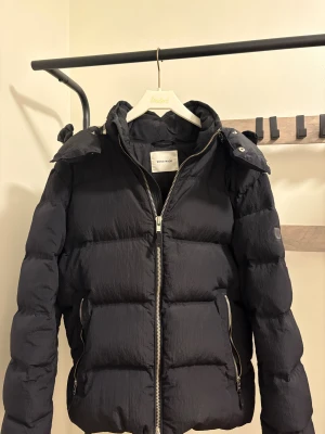Svart pufferjacka från Wood Wood - Svart vinterjacka från Wood Wood i storlek S. Jackan har en avtagbar huva. Höger jackficka är lagad pga av hål. Köpt för 5300. Hör av er om funderingar!