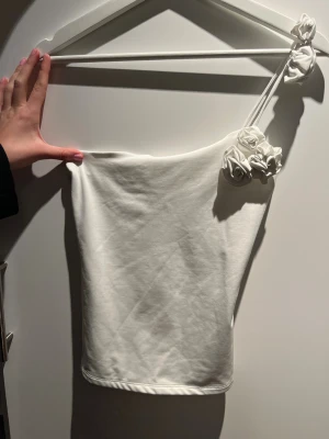 Vit oneshoulder topp med blommor - Säljer en vit oneshoulder topp med axelbandslös design och dekorativa tygblommor på axelbandet. Toppens form är tight och den har en stilren, modern look.