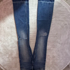 Blå bootcut jeans från H&M - Säljer ett par blå bootcut jeans från H&M med låg midja och justerbar midja. Jeansen har slitna detaljer och snygga slitningar på benen, klassisk femficksdesign och knappstängning. Perfekt för dig som gillar en avslappnad och trendig look.