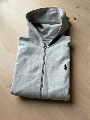 Grå hoodie från Polo Ralph Lauren - Snygg grå hoodie från Polo Ralph Lauren med dragkedja och huva. Klassisk svart logga på bröstet och två fickor framtill. Tillverkad i mjuk bomullsmix, perfekt för chill dagar. Passformen är normal och ärmarna är långa.