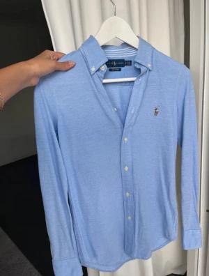 Blå skjorta från Ralph Lauren - Klassisk ljusblå skjorta från Ralph Lauren med button down-krage och broderad logga på bröstet. Skjortan har långa ärmar, vita knappar och är tillverkad i mjuk bomull. Köpt för 2000 kr
