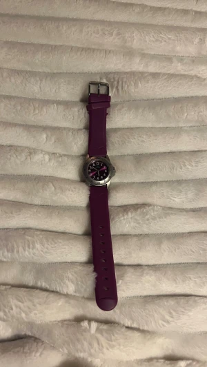 Lila klocka från GUL med silvrigt ur - Säljer en snygg klocka från GUL med lila silikonarmband och rund silvrig boett. Urtavlan är lila med vita siffror och detaljer i silver. Klockan har klassisk spänne och sportig design, perfekt för dig som gillar färg och vill sticka ut. OBS!! Batteriet behöver dock bytas!