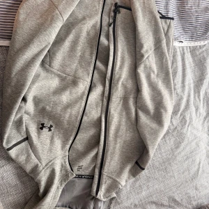 Grå zip-hoodie från Under Armour - Snygg grå zip-hoodie från Under Armour med svart logga på bröstet och svarta detaljer vid dragkedjan. Jackan har huva, långa ärmar och är tillverkad i mjuk bomullsblandning. Perfekt för träning eller chill dagar.
