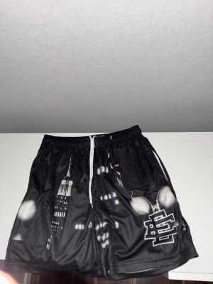 Svarta shorts med stadsmotiv och snörning - Snygga svarta shorts med coolt stadsmotiv i vitt och grått, samt en vit snörning i midjan. Materialet är troligtvis polyester och de har en loose passform. Perfekta för dig som gillar streetstyle och grafiska prints.