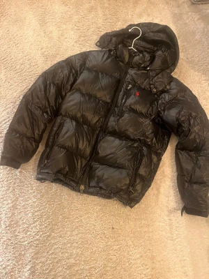 Svart pufferjacka med huva från Polo Ralph Lauren - Säljer en svart pufferjacka från Polo Ralph Lauren med huva och flera dragkedjefickor. Jackan har en röd Polo-logga på bröstet, quiltad design och justerbar nederkant med snörning. Perfekt för kalla dagar och riktigt snygg streetstil.