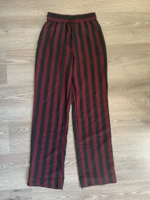 Randiga byxor från Monki XXS - Snygga byxor från Monki i storlek XXS med breda vertikala ränder i vinrött och svart. Byxorna har resår och snörning i midjan samt en bakficka. De är raka i modellen och tillverkade i mjukt bomullstyg, perfekta för en chill och trendig look.
