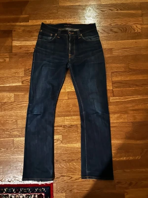 Mörkblå jeans från Nudie Jeans - Snygga mörkblå jeans från Nudie Jeans med klassisk femficksdesign och raka ben. Jeansen har kontrastsömmar och läderpatch med orange logga bak i midjan. Perfekta för dig som gillar stilrena och tidlösa jeans i kraftig denim. Ganska slim i passformen hör av er vid frågor!