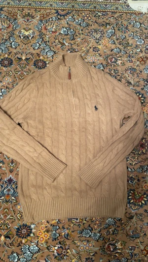 Polo Ralph Lauren Tröja - Snygg beige stickad tröja från Polo Ralph Lauren med klassiskt flätmönster och broderad logga på bröstet. Tröjan har hög krage med dragkedja och ribbade muddar. Perfekt för dig som gillar stilrena och tidlösa plagg. Står M men sitter som S!