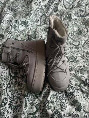 Grå knutna boots med platt sula - Säljer ett par grå knutna boots med platt sula och rund tå. Skorna har snörning framtill och är fodrade med mjukt syntetmaterial på insidan för extra värme. Perfekta för kalla dagar och har en chunky look som sticker ut. Ett billigare alternativ för inuikii skor! Super fina och super sköna 🤍 Bara använda nån enstaka gång!