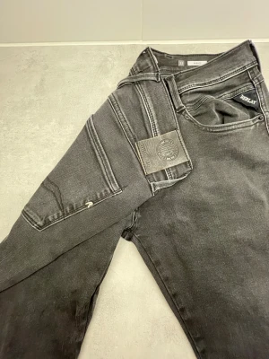 Replay Anbass svarta jeans, storlek 32 - Svarta Replay Anbass jeans med snyggt tvättad look och klassiska fem fickor. Jeansen har raka ben, normal passform och detaljerade sömmar på bakfickorna. Materialet är stretchigt jeans för skön komfort och stil.
