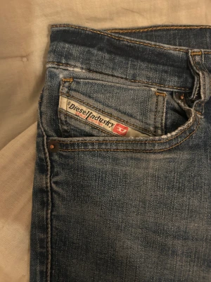 Blå jeans från Diesel, straight fit - Säljer ett par blå jeans från Diesel med klassisk femficksdesign och snygga slitningar framtill. Jeansen har raka ben och normal passform, med orangea kontrastsömmar och Diesel Industry-märke vid fickan. Materialet är bomull, polyester och elastan för skön stretch.