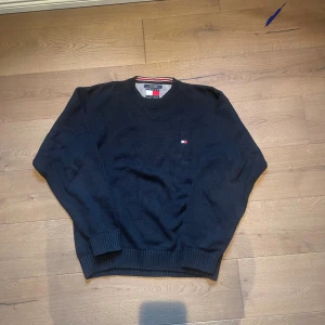 Mörkblå stickad tröja Tommy Hilfiger - Snygg mörkblå stickad tröja från Tommy Hilfiger med klassisk rund hals och diskret logga broderad på bröstet. Tröjan är gjord i 100% bomull och har ribbade muddar vid ärmslut och nederkant. Perfekt för dig som gillar stilrena och tidlösa plagg.