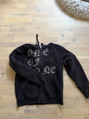 One of one  - Svart hoodie med dragkedja och huva, dekorerad med stora bokstäver i silvrigt tryck som bildar texten 'ONE OF ONE' på framsidan. Tröjan har långa ärmar och två fickor framtill. Perfekt för dig som gillar streetwear och vill sticka ut. 