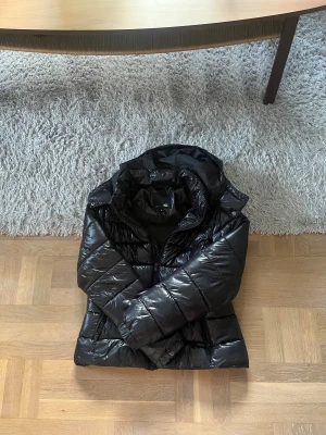 Svart pufferjacka från lager 157, M - Säljer en svart, glansig pufferjacka från lager 157 i storlek M. Jackan har huva, dragkedjor framtill och på fickorna samt quiltad design. Perfekt för kalla dagar och riktigt snygg streetstil.