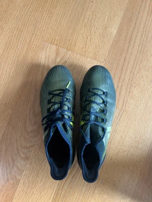 Adidas svarta och gula fotbollsskor - Säljer ett par Adidas fotbollsskor med svart och gul färg, täckt av små prickar och med Adidas-logga på sidan. Skorna har snörning och platt sula med fasta dobbar för gräsplan. Ovandelen är i syntetmaterial och har en smal, sportig form. Storleken är 41 1/3 men funkar för både strl 41 och 42