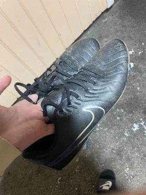 Svarta Nike fotbollsskor med blå detaljer - Säljer ett par svarta Nike fotbollsskor med snygga blå och vita detaljer. Skorna har snörning och är tillverkade i syntetmaterial med en rund tå och platt sula med dobbar för bra grepp på planen. Perfekt för dig som vill ha stil och funktion på träningen eller matchen.