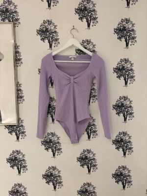Ljuslila ribbad långärmad body - Säljer en ljuslila body från H&M med långärmade och ribbad struktur. Toppen har en snygg v-ringad hals och en twistad detalj framtill. Materialet är mjukt och stretchigt, perfekt för att skapa en clean och trendig look.