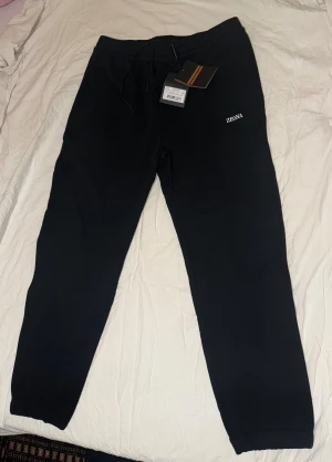 Svarta mjukisbyxor från Zegna - Svarta mjukisbyxor från Zegna med broderad logga på vänster ben. Byxorna har snörning i midjan och elastiska muddar vid bensluten. Tillverkade i mjukt bomullsmaterial för extra komfort. Perfekta för chill eller träning.