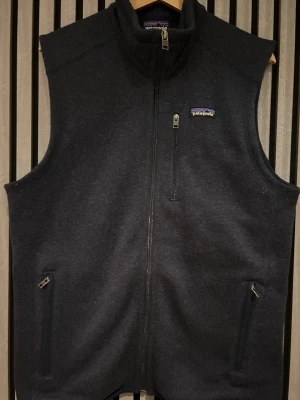 Mörkblå fleeceväst från Patagonia - Snygg mörkblå fleeceväst från Patagonia med hög krage och tre praktiska fickor med dragkedja. Västen har en stilren look och är perfekt för lager-på-lager. Mjuk insida och diskret logga på bröstet. Passar dig som gillar outdoor och chill stil. Vänsten är använd relativt mycket men är i fint skick och inga defekter