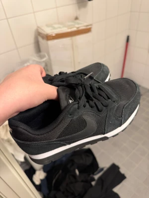 Svarta Nike sneakers med snörning - Snygga svarta Nike sneakers med klassisk swoosh på sidan. Skorna har mesh och mockadetaljer, platt sula och rund tå. Perfekta för dig som gillar stilrena och sportiga skor. Svarta skosnören och vit kontrastsula ger en clean look.