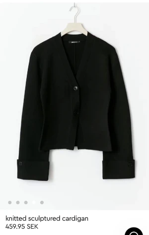 Svart kofta från Gina Tricot - Stilren svart kofta från Gina Tricot med v-ringad hals och två knappar framtill. Koftan har långa ärmar med breda muddar och knappdetaljer vid ärmslut. Den är fin stickad och har en skulpturerad, lite boxig passform som ger en cool vibe.