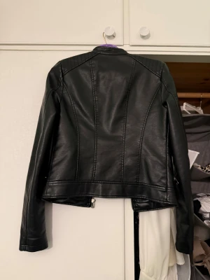 Svart skinnjacka från Vero Moda - Snygg svart skinnjacka från Vero Moda med raka linjer och coola detaljer som dragkedjor vid fickorna och framtill. Jackan har quiltade axelpartier och en klassisk krage. Perfekt för dig som vill ha en edgy stil och gillar att sticka ut.