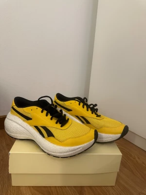 Reebok gula sneakers med plattform - Säljer ett par snygga Reebok sneakers i starkt gul färg med svarta detaljer och logga. Skorna har svart snörning, rund tå och en rejäl vit plattformssula som ger extra höjd och cool vibe. Ovandelen är i mesh och syntet för bra andning och komfort.