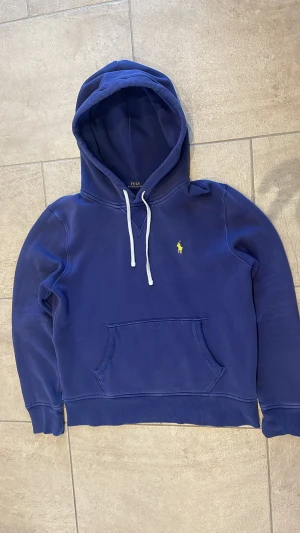Blå hoodie från Polo Ralph Lauren - Snygg marinblå hoodie från Polo Ralph Lauren med klassisk gul logga på bröstet. Tröjan har huva med vita snören, magficka och ribbade muddar. Perfekt för chill dagar och enkel att matcha med jeans eller joggers.   Tröjan liten i storleken och passar någon som bär S bättre
