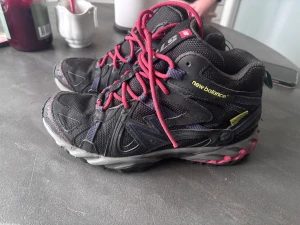 Svarta New Balance sneakers strl 37.5 - Säljer ett par svarta New Balance sneakers med coola rosa snören och detaljer i lila och grått. Skorna har en robust och sportig design med mesh och syntetmaterial, perfekt för dig som gillar streetstyle och komfort. Rund tå och platt sula för skön känsla.