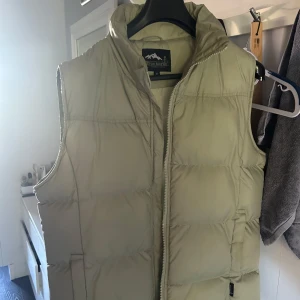 Grön dunväst från True North - Säljer en grön dunväst från True North med quiltad design och hög krage. Västen har dragkedja framtill och två sidofickor. Perfekt för lager-på-lager och chill höstdagar. Enkel och clean look som funkar till många stilar.