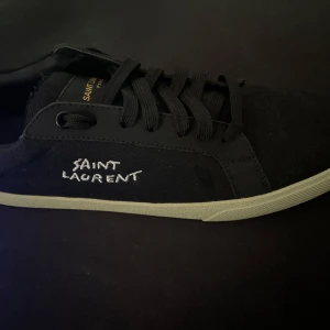 Svarta Saint Laurent sneakers - Snygga svarta sneakers från Saint Laurent med vit broderad logga på sidan och gulddetaljer på plösen. Skorna har svart snörning, vit platt sula och är tillverkade i canvas med detaljer i skinn. Perfekta för dig som gillar stilrena och exklusiva sneakers. Storlek:44