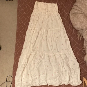 Vit blommig långkjol från 157 - Superfin långkjol i vitt från 157 med små blommiga detaljer i pastellfärger. Kjolen har smock i midjan och ett luftigt, flowy tyg som ger en romantisk vibe. Perfekt för dig som gillar en feminin och avslappnad stil.