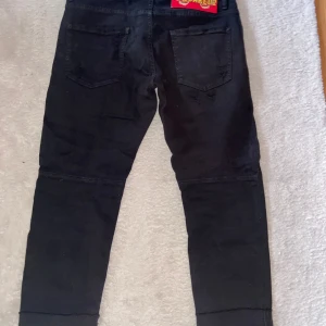 Svarta jeans från Dsquared2, storlek 46 - Säljer ett par svarta jeans från Dsquared2 i storlek 46. Jeansen har slitna detaljer framtill, klassisk femficksmodell och snygg Dsquared2-patch med tigrar bak. Materialet är bomull med en skön jeanskänsla och passformen är slim.