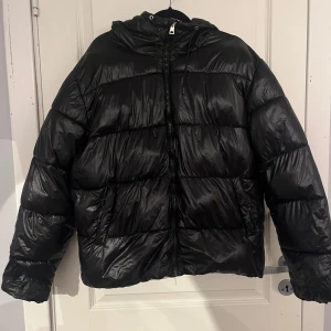 Svart pufferjacka från 157 - Säljer en svart pufferjacka från 157 med huva och dragkedja framtill. Jackan är quiltad och har en glansig finish, perfekt för kalla dagar. Modellen är oversized och har snörning i huvan för extra komfort. Riktigt skön och trendig jacka för vintern! Sparsamt använd🩵