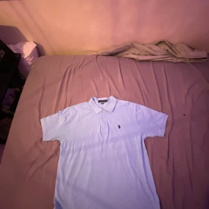 Ljusblå polo t-shirt från U.S. Polo Assn. - Fräsch ljusblå polo t-shirt från U.S. Polo Assn. med klassisk krage och knappar framtill. T-shirten har korta ärmar och en broderad logga på bröstet. Materialet är mjuk bomull som känns skönt mot huden. Perfekt för dig som gillar stilren och enkel design.