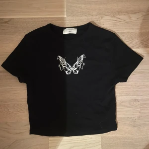 Svart croppad t-shirt med fjäril - Svart magtröja från Chiquelle med vit fjärilstryck på bröstet. Säljer då den är lite för liten för mig. Köptes för länge sedan men använt bara en gång.