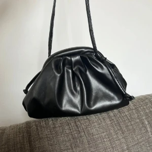 Clutch bag  - Snygg svart axelväska i skinnimitation med mjuk, puffig form och dragsko upptill. Väskan har en minimalistisk look med släta ytor och smala axelband. Perfekt för dig som gillar stilrena accessoarer med en modern twist.