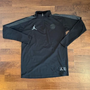 Svart PSG x Jordan half zip tröja - Svart långärmad tröja från Jordan x PSG med halv dragkedja och högt krage. Tröjan har grå detaljer på axlarna, PSG-logga och Jumpman-logga på bröstet samt patch nertill. Perfekt för dig som gillar sportig och clean stil. Storleken är L men passar dig som brukar ha M i tröjor. 