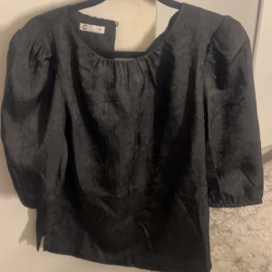 Svart blus med puffärm från C - Stilren svart blus  med rund halsringning och  puffärmar i trekvarts längd.  ett glansigt material ,en elegant look.