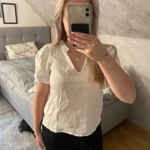 Vit blus med puffärm från Desires - Supersöt vit blus från Desires med puffiga korta ärmar, v-ringning och liten volangkant vid halsen. Tillverkad i mjuk bomull med lätt skrynklig struktur. Perfekt för en fräsch och stilren look.