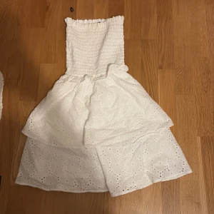 Vit axelbandslös sommarklänning - Supersöt vit axelbandslös mini klänning med smockad överdel och broderade blommor på kjolen. Klänningen har en kort, voluminös kjol i två lager och är perfekt för varma dagar. Tillverkad i bomull för en luftig känsla.