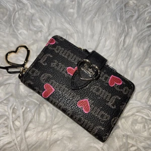 Juicy Couture plånbok med hjärtan - Supersöt plånbok från Juicy Couture i svart med rosa hjärtan och logga i mönstret. Plånboken har ett hjärtformat spänne i metall, flera kortfack, myntficka med dragkedja och en beige insida. Perfekt accessoar för dig som gillar detaljer och trendig design.